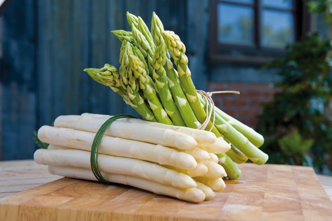 Bund weißer Spargel auf einem Holzbrett. Auf den weißen Spargel ist ein Bund grüner Spargel gelehnt.
