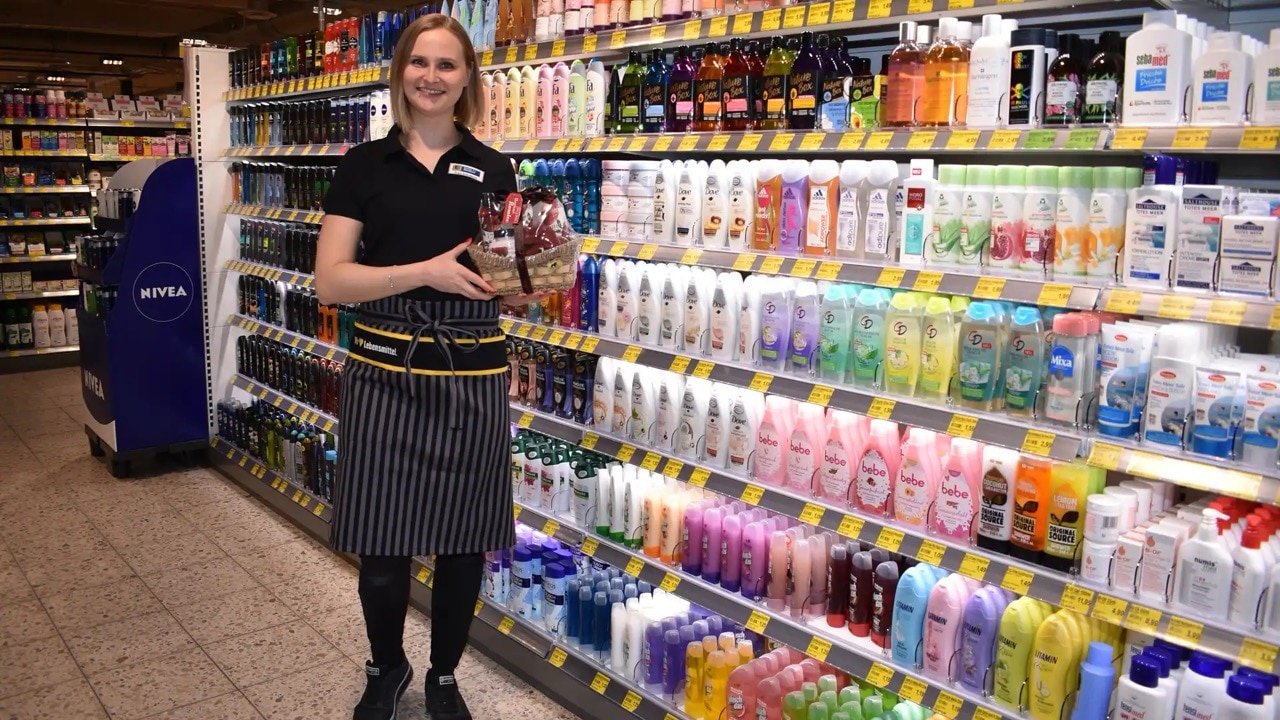 Eine Frau in schwarzer EDEKA-Kleidung steht vor einem Drogerieregal und hält einen kleinen Präsentkorb.