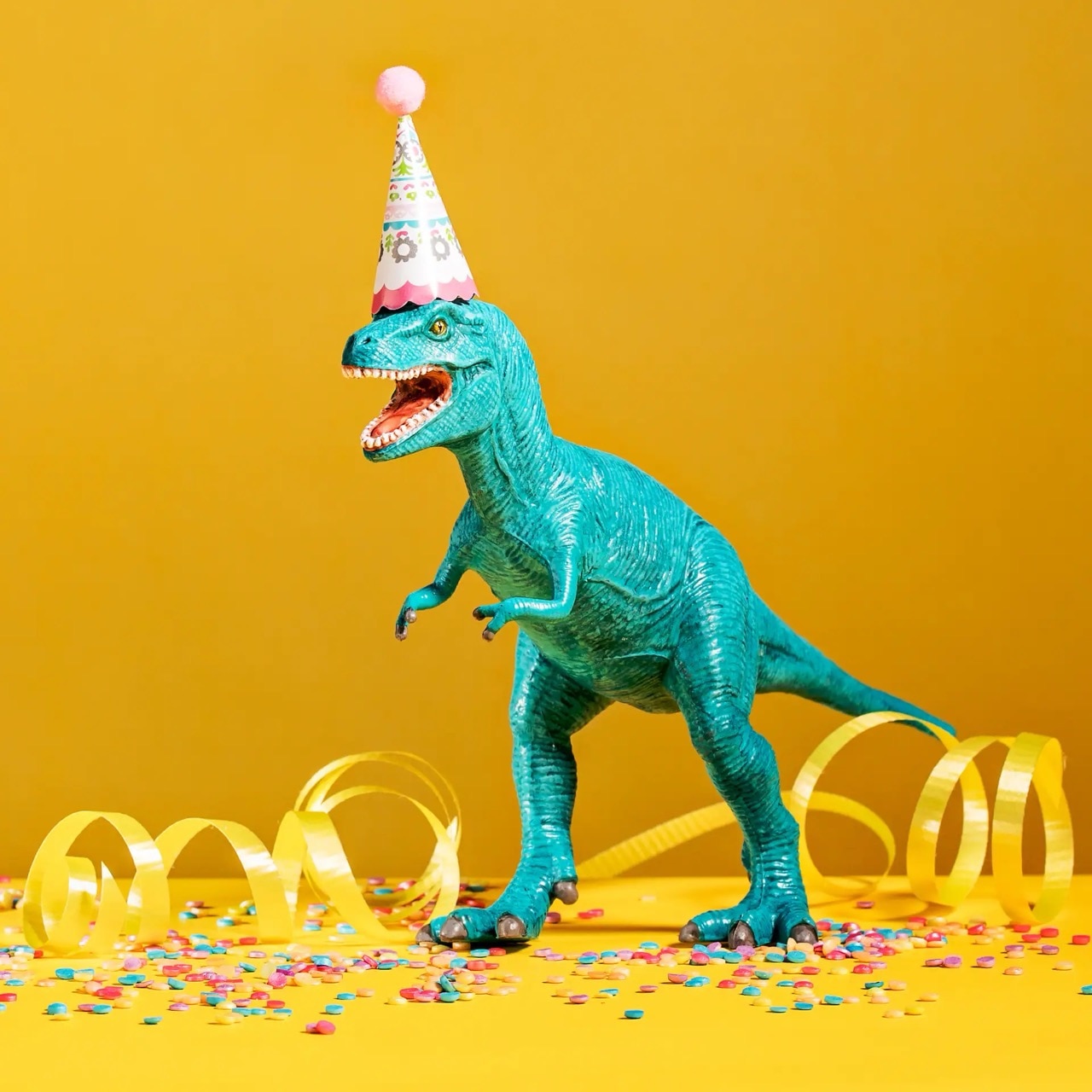 Grüner Plastic-Dino mit Partyhut. Auf dem Boden Konfetti und Luftschlangen.