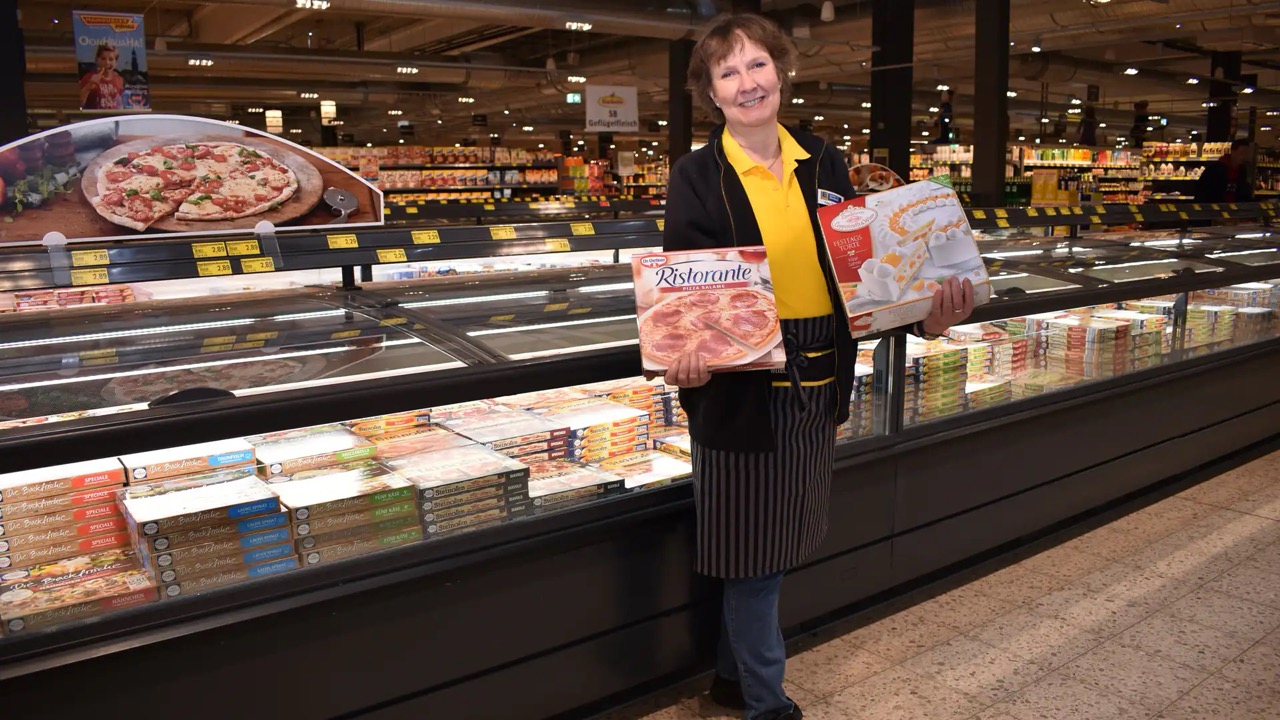 Vor einer Tiefkühltruhe steht eine Frau, die in ihren Händen eine Tiefkühl-Pizza sowie TK-Torte hält. Sie trägt schwarz-gelbe EDEKA-Kleidung.
