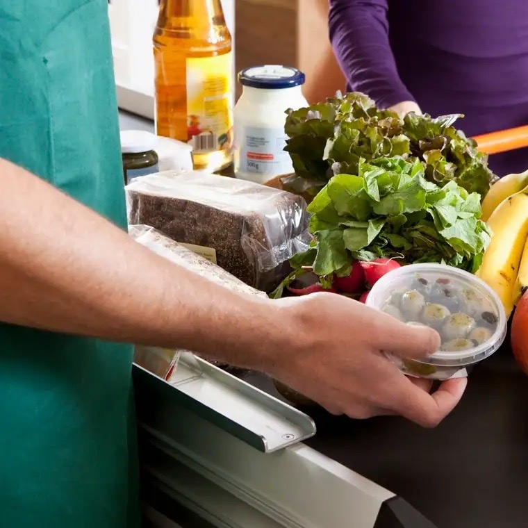 Eine Hand hält einen Plastic-Behälter mit Oliven über ein Kassenband.