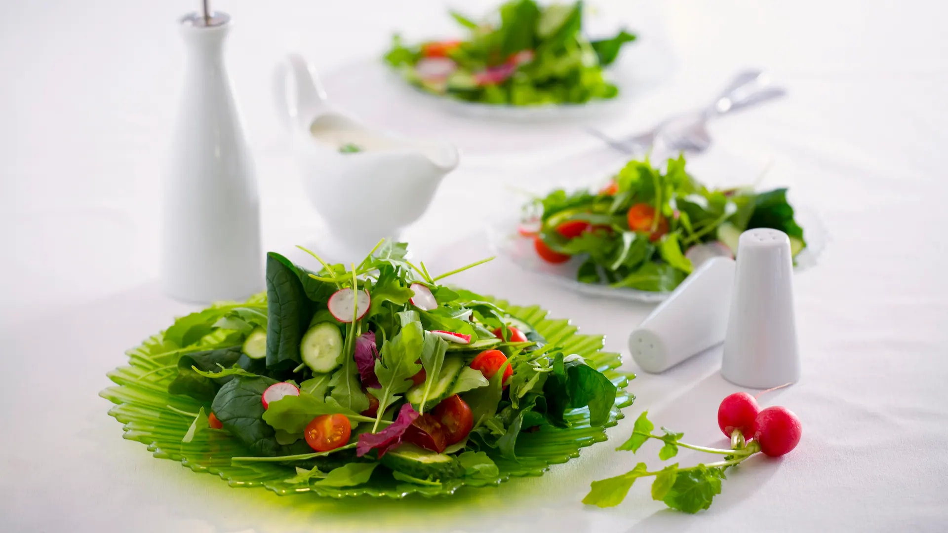 Grüner Teller mit Salat auf einer weißen Tischdecke. Im unscharfen Hintergrund stehen zwei weitere Teller Salat.