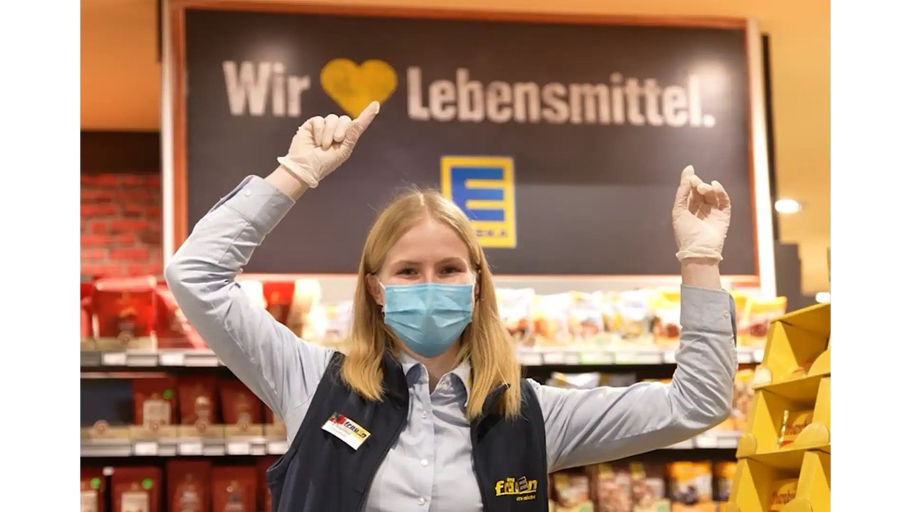 Eine Frau in EDEKA-Kleidung streckt beide Zeigefinger nach oben. Über ihr steht auf einem Schild: Wir lieben Lebensmittel.