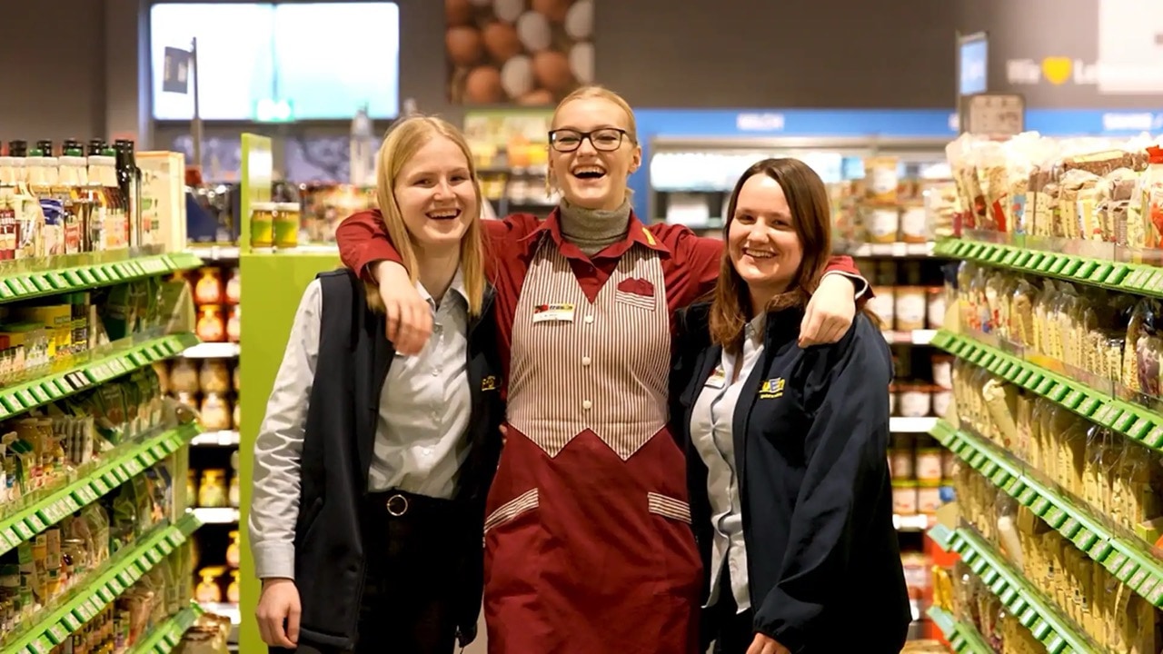 Drei Frauen in EDEKA-Kleidung stehen in einem Gang mit Regalen. Die Frau in der Mitte hat ihre Arme um die Schultern der beiden anderen gelegt.