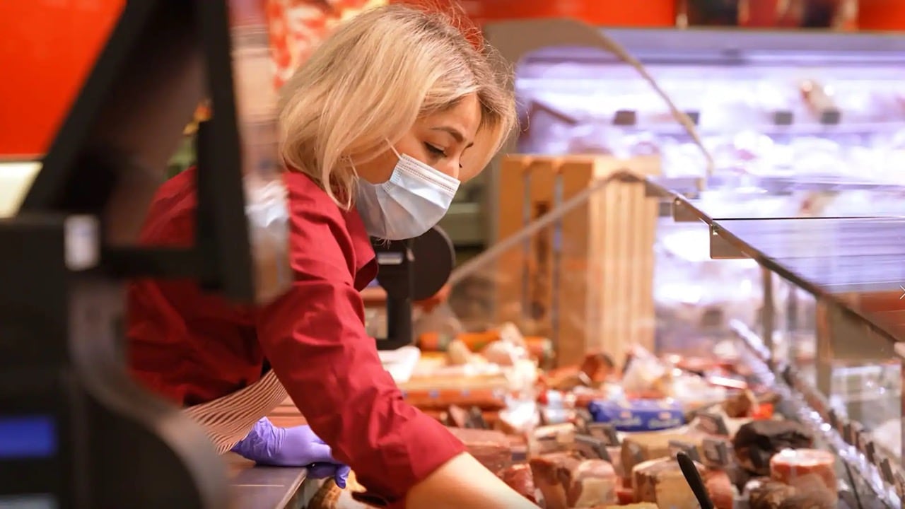 Eine Frau mit rotem Hemd und Schürze greift über die Frischetheke. Sie trägt einen Mund-Nasenschutz.