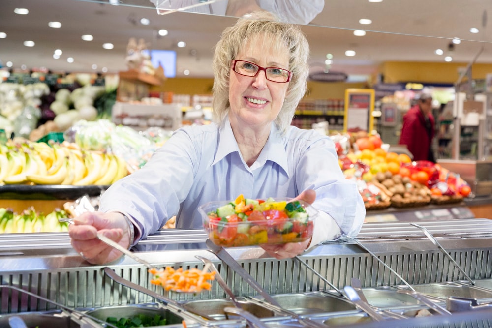 Eine Frau füllt einen transparenten Einwegbehälter mit Produkten aus der Salatbar.