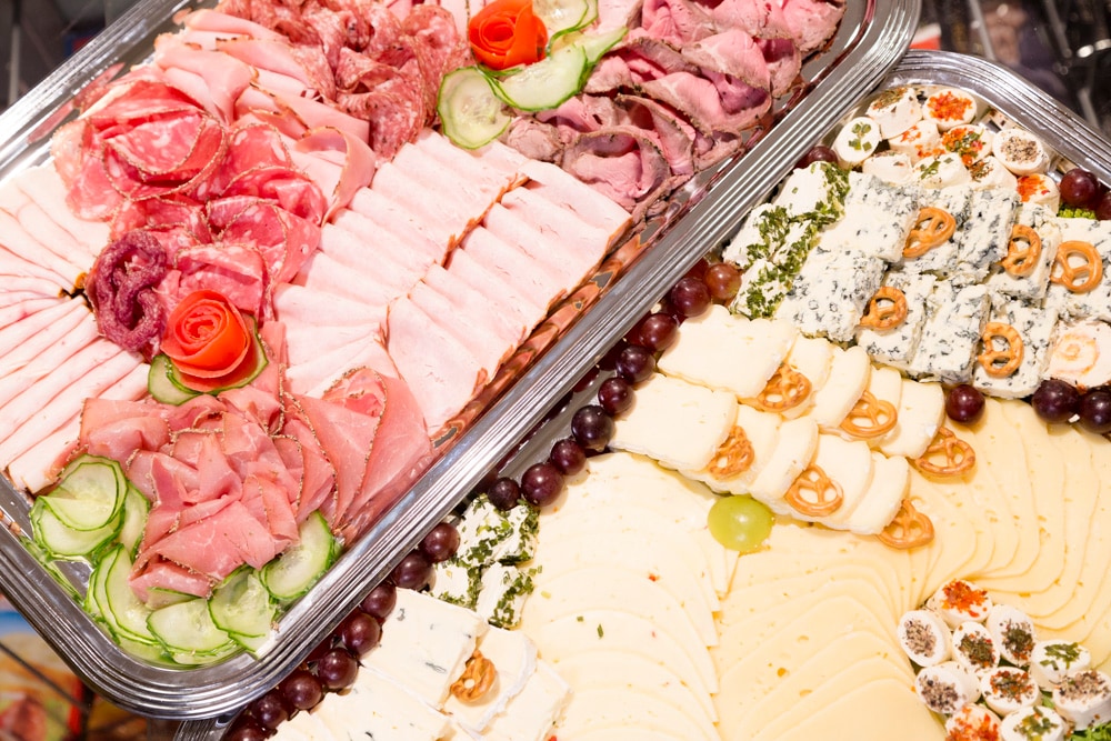 Zwei kalte Platten, eine mit Wurstaufschnitt und eine mit Käse. Als Deko dienen Gurkenscheiben, Weintrauben und Salzbrezeln.