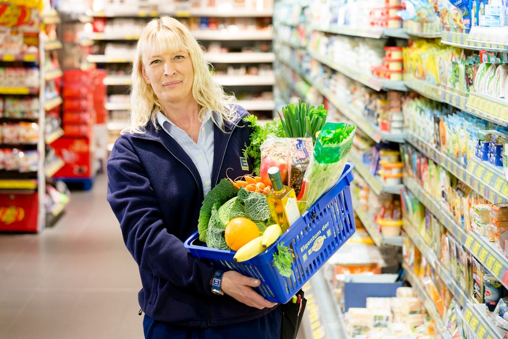 Eine Frau in blauem EDEKA-Fleece-Pullover hält einen gefüllten Einkaufskorb hoch.