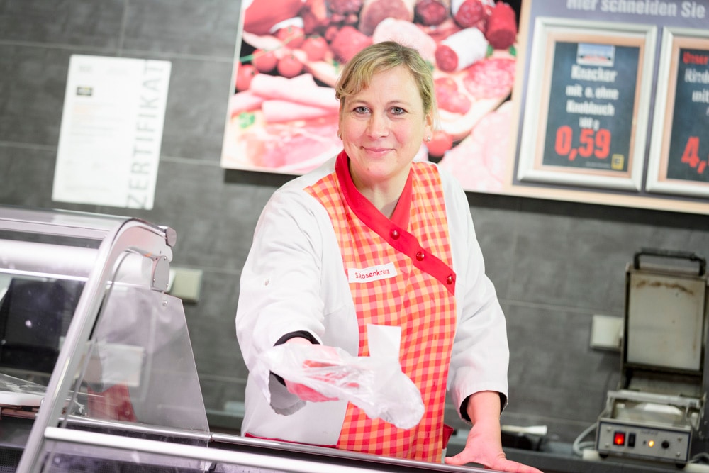 Eine lächelnde Frau steht hinter der Fleischtheke und streckt ihre Hand mit einer verpackten Ware aus.