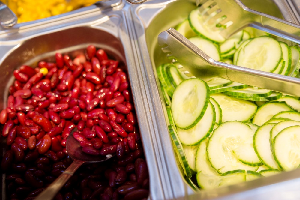 Zwei Edelstahl-Behälter der Salatbar. Einer mit Kidney-Bohnen, einer mit Gurkenscheiben.