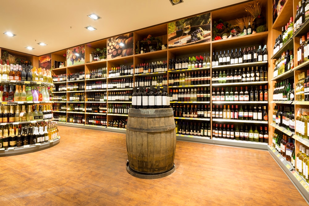 Weinregale im Supermarkt. Davor eine Holztonne, auf der Weinflaschen stehen.