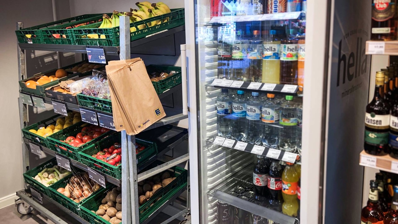 Neben einem Obst- und Gemüseständer steht ein Kühlschrank mit Getränken.