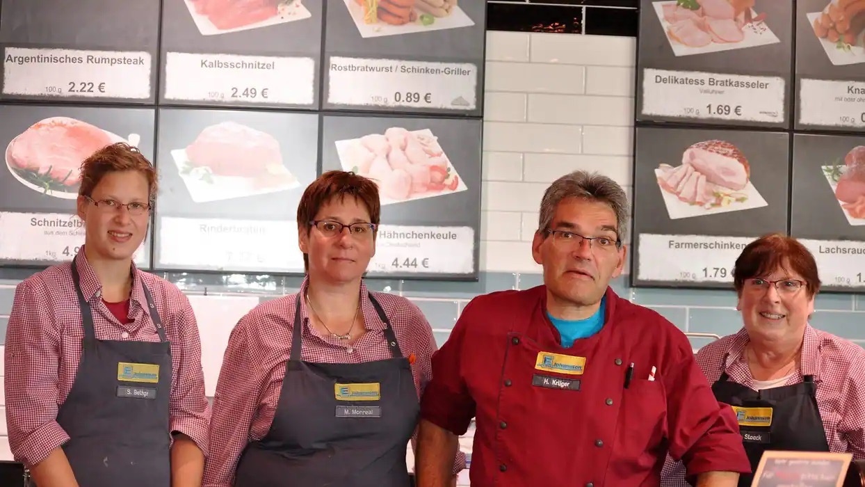 Drei Frauen und ein Mann stehen hinter Fleischtheke. Die Frauen tragen rot-weiß-karierte Hemden unter einer dunkelgrauen Schürze. Der Mann trägt eine rote Fleischerjacke.