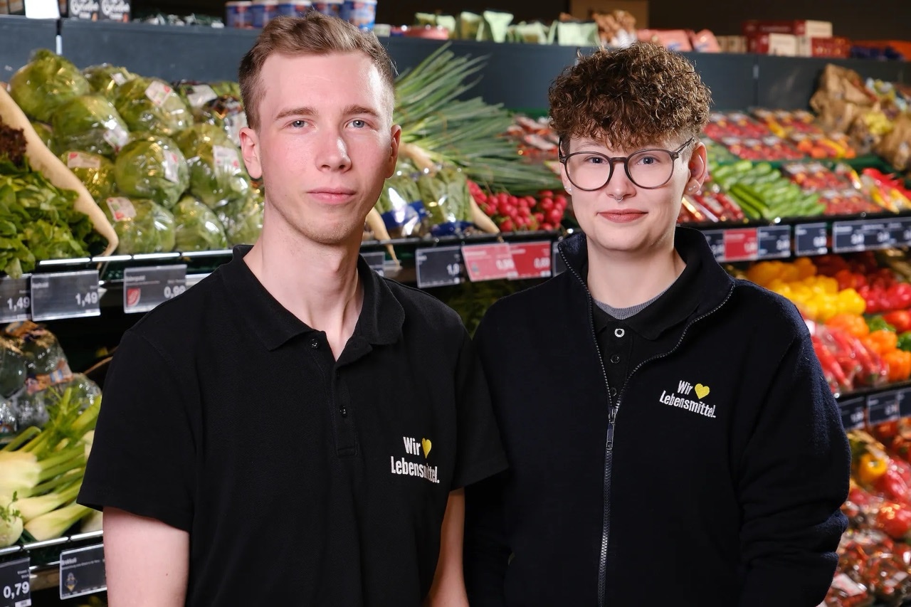 Zwei junge Menschen in schwarzer EDEKA-Kleidung vor dem Gemüseregal.