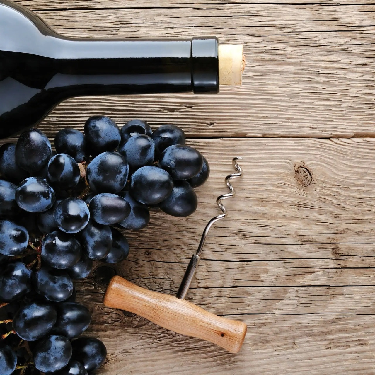 Auf einem Holztisch liegen eine Flasche Rotwein, blaue Trauben und ein Korkenzieher.