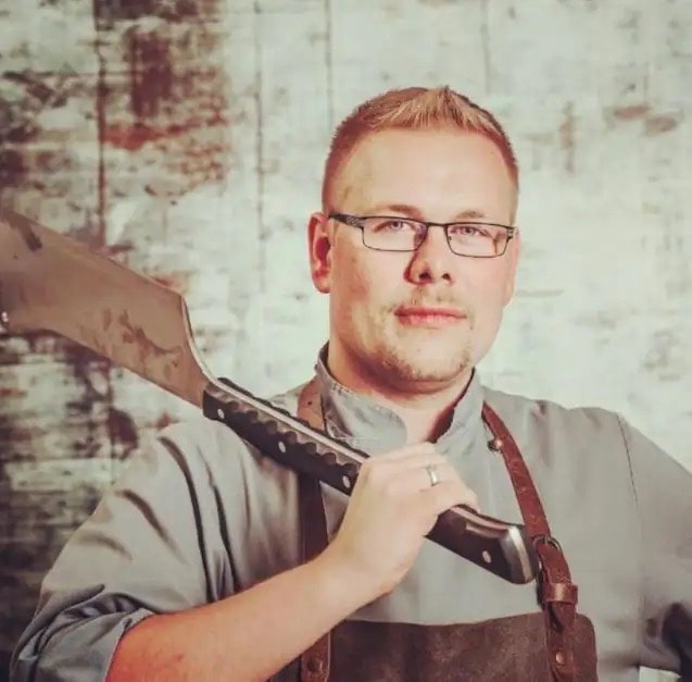 Ein Mann mit Schürze und grauem Fleischhemd hält ein großes Messer über seiner rechten Schulter.