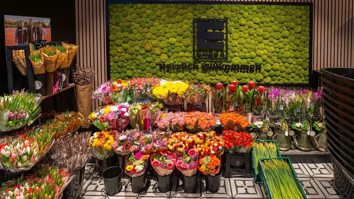 Verpackte Blumengestecke in schwarzen Eimern. Dahinter eine grüne Mooswand mit schwarzem EDEKA-Logo und der Aufschrift Herzlich WIllkommen