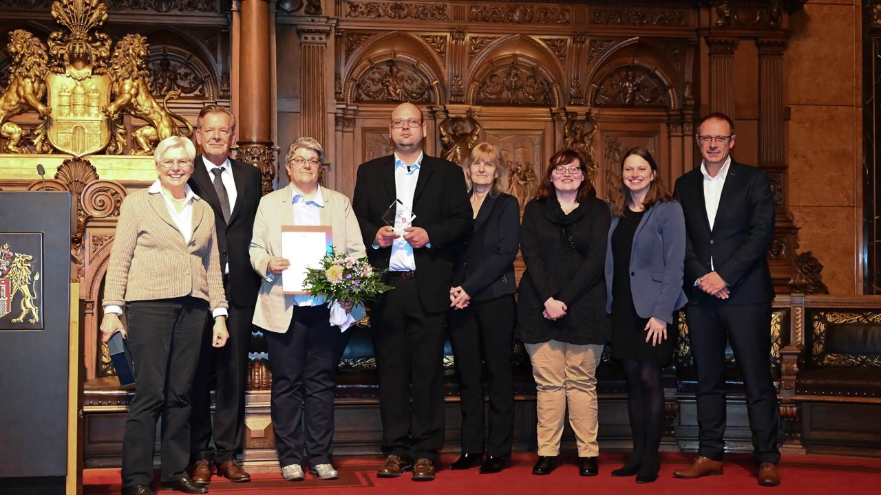 Acht Personen in Abendkleidung auf einem roten Teppich. Eine Frau hält eine Urkunde, der Mann neben ihr eine Auszeichnung.