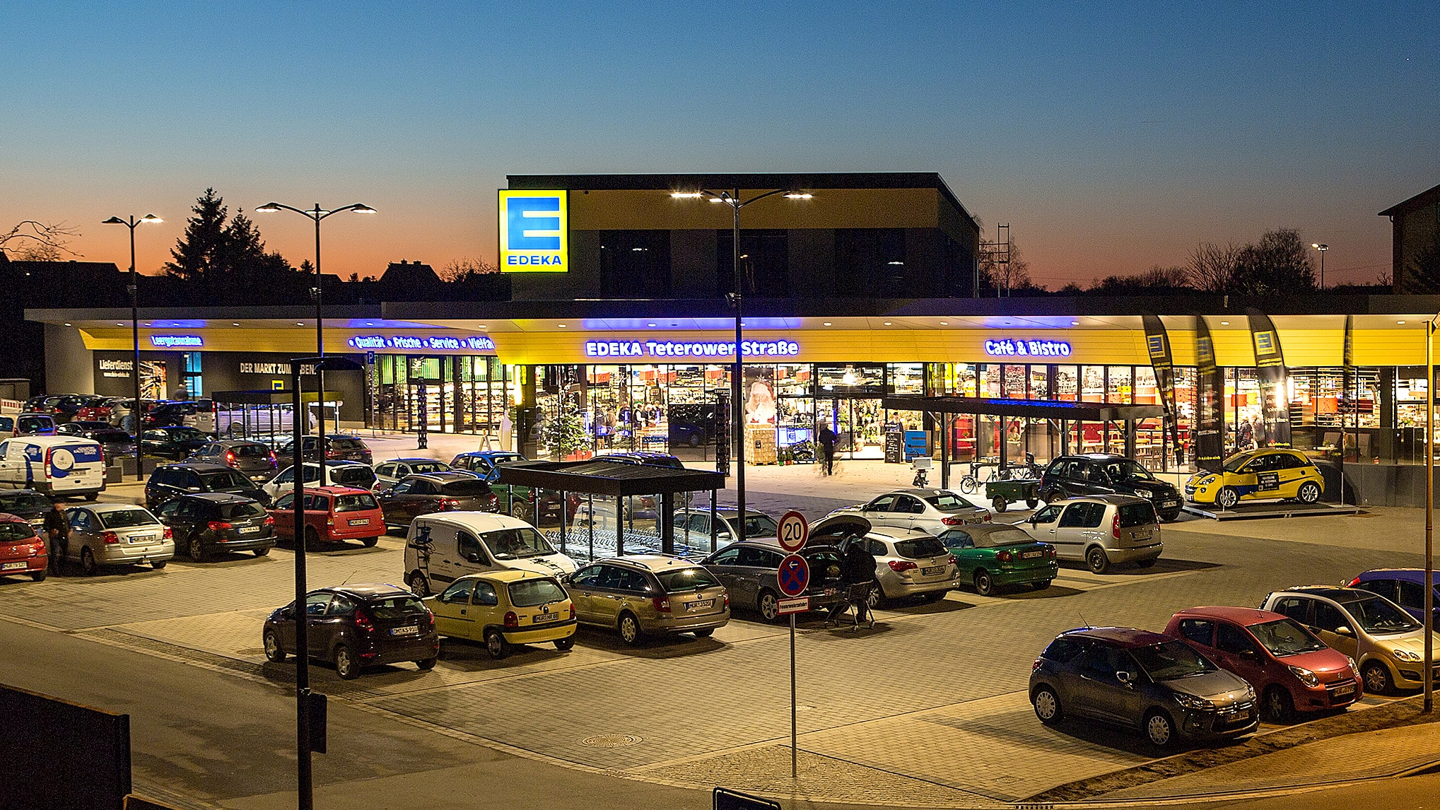 Beleuchteter EDEKA-Eingang und voller Parkplatz im Sonnenuntergang.