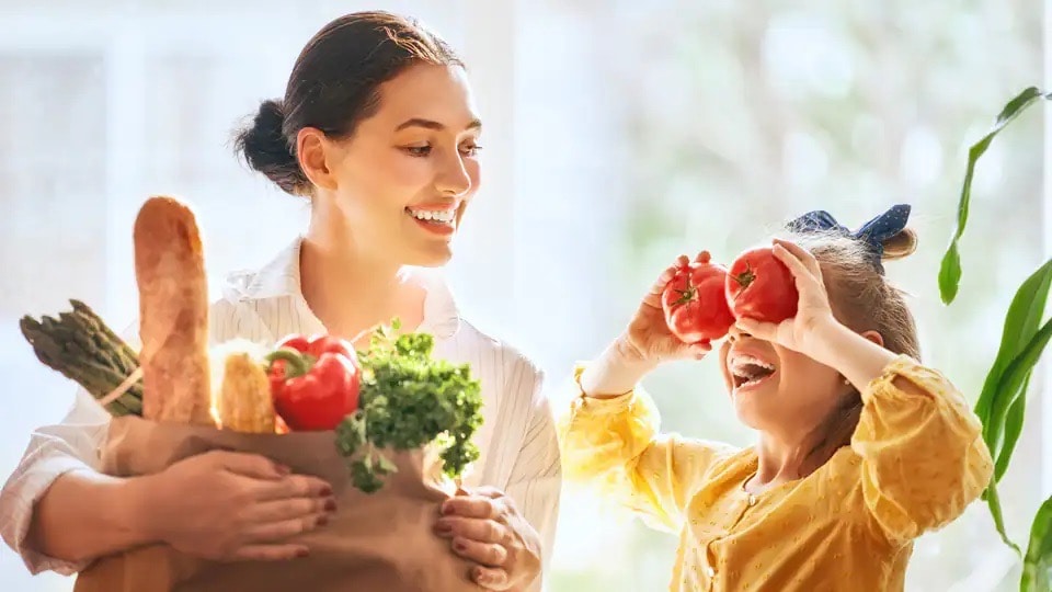Lachende Frau hält eine Papiertüte mit Lebensmitteln. Neben ihr ein junges Mädchen, das sich zwei rote Paprika vor die Augen hält.