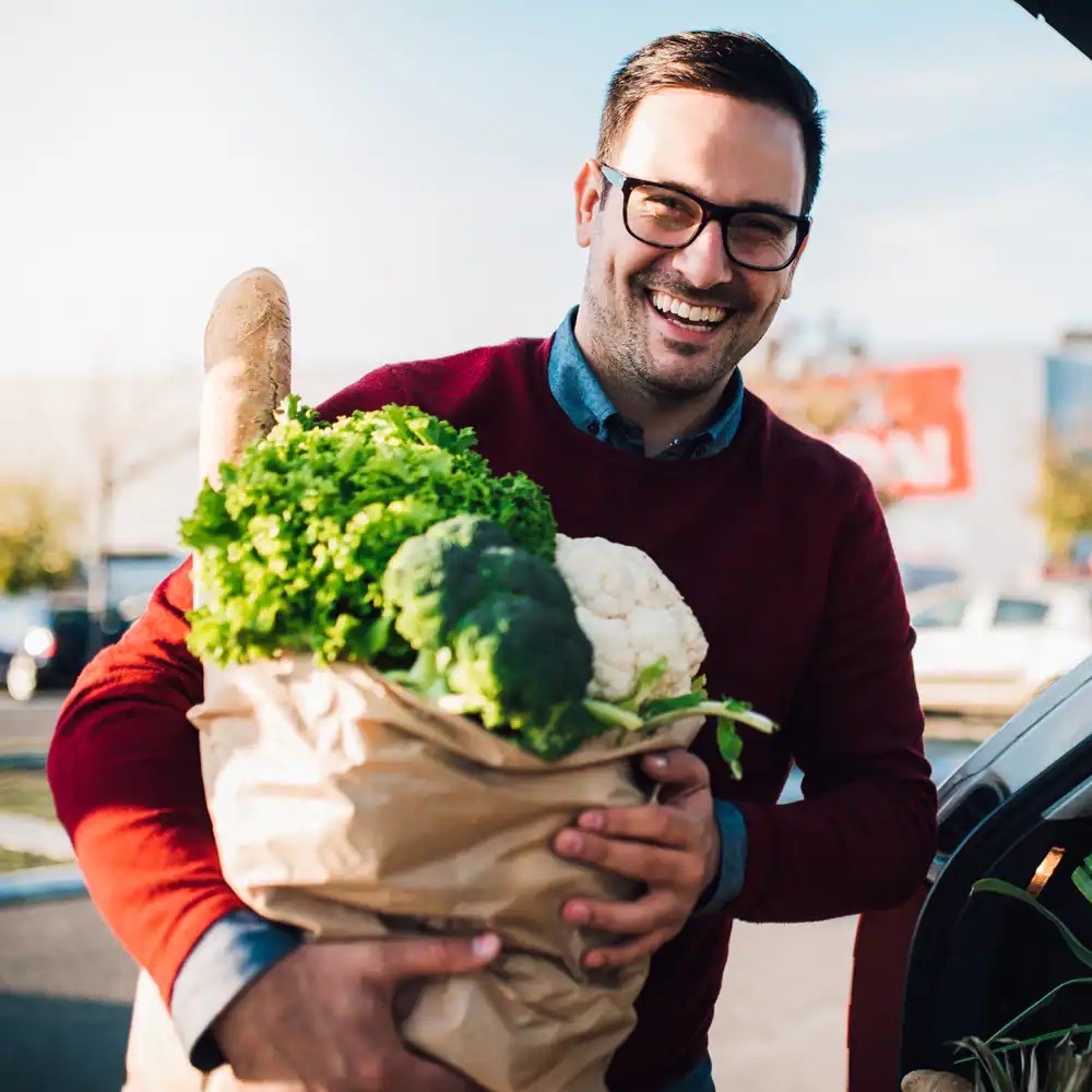 Lachender Mann steht an einem Auto und hält eine Papiertüte mit Lebensmitteln.