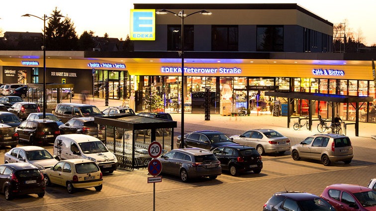 Beleuchteter EDEKA-Eingang und voller Parkplatz im Sonnenuntergang.