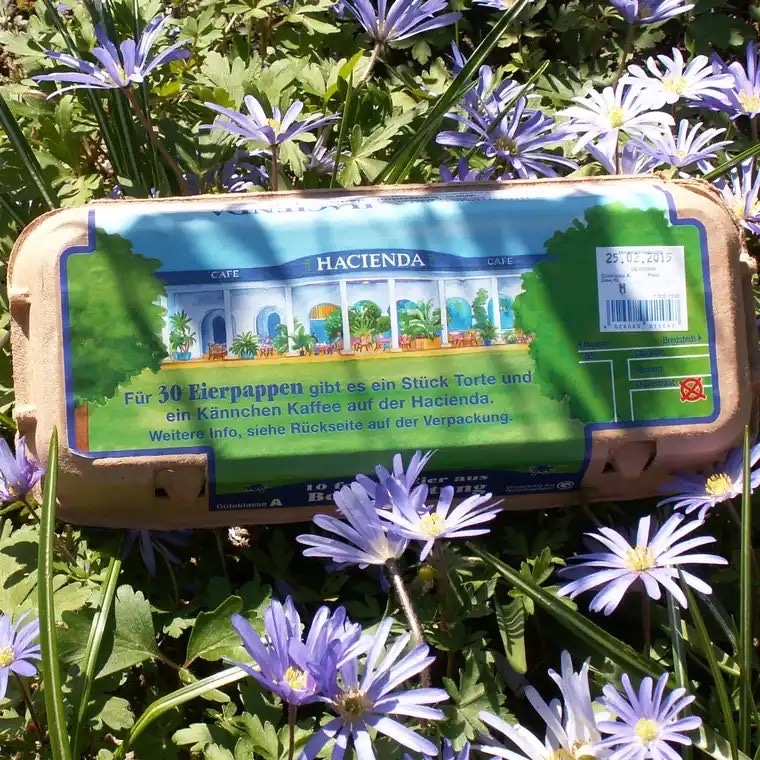 Eierkarton liegt auf lilanen Blumen. Aufschrift: Für 30 Eierpappen gibt es ein Stück Torte und ein Kännchen Kaffee auf der Hacienda. Weitere Info, siehe Rückseite auf der Verpackung.