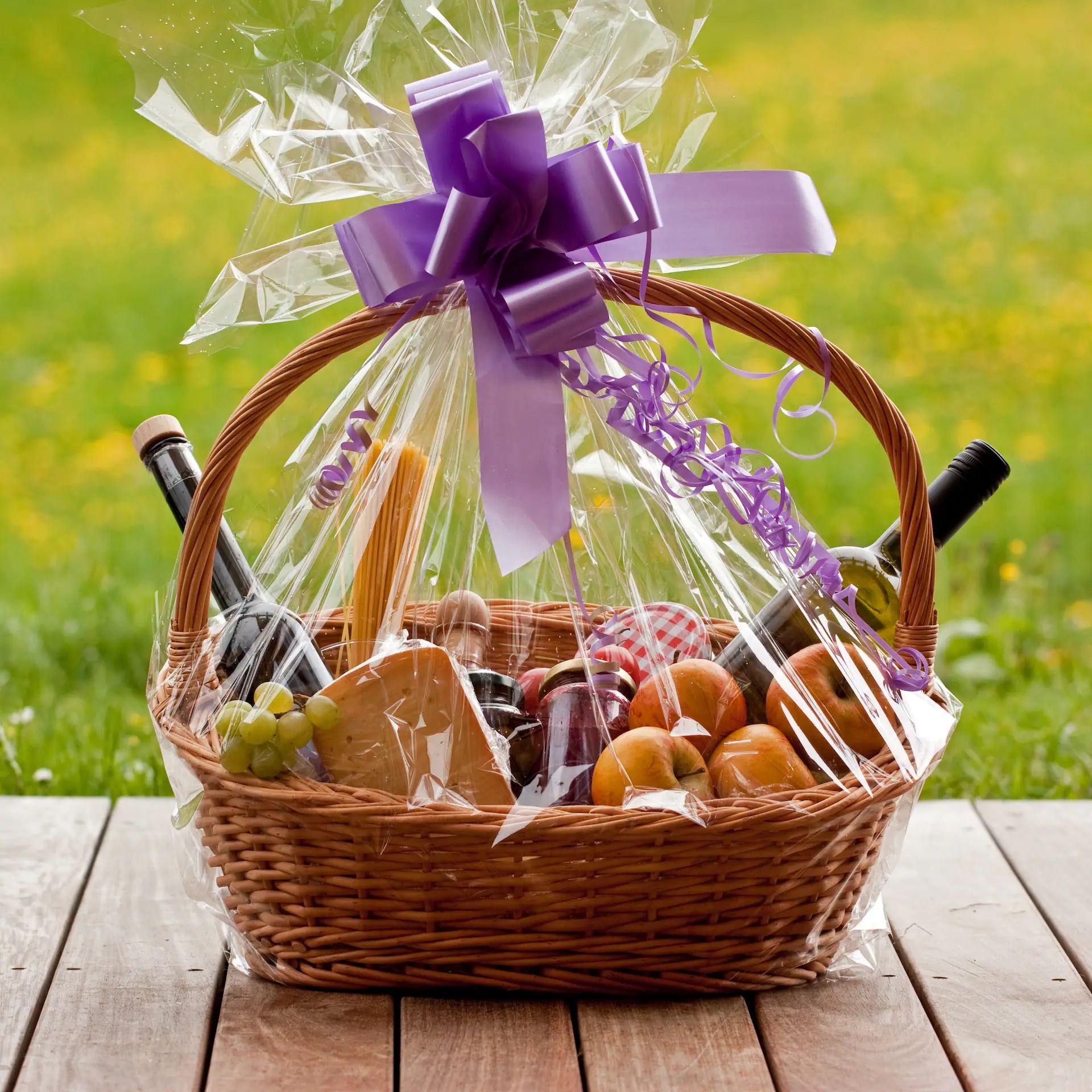 Präsentkorb mit lilaner Schleife. Unter der Klarsichtfolie liegen Äpfel, Marmelade, Käse, Weintrauben, Öl und eine Flasche Wein.