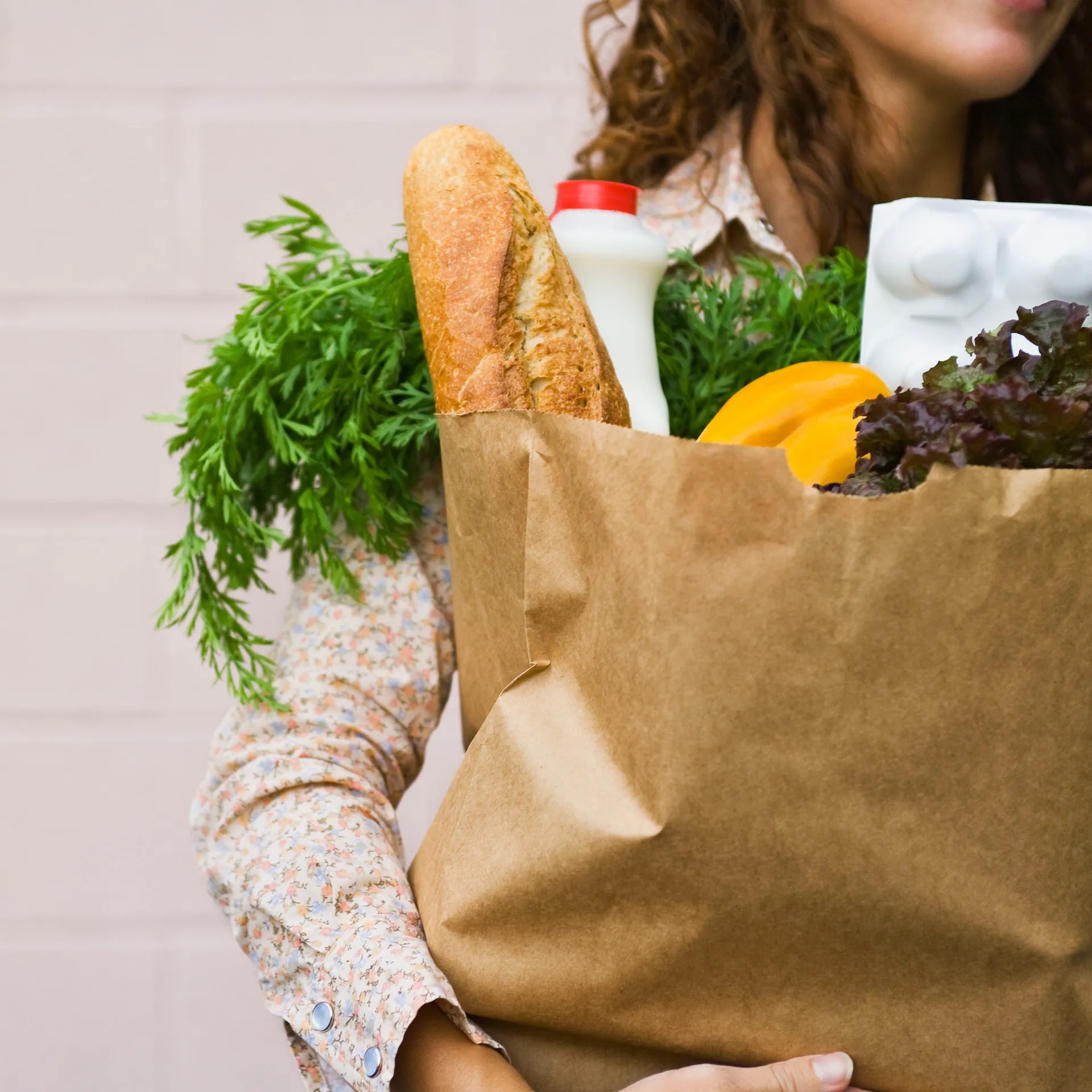 Eine Frau hält eine Einkaufstüte aus Papier im Arm. Es ragen ein Baguette, Kräuter, Paprika und Salat heraus.