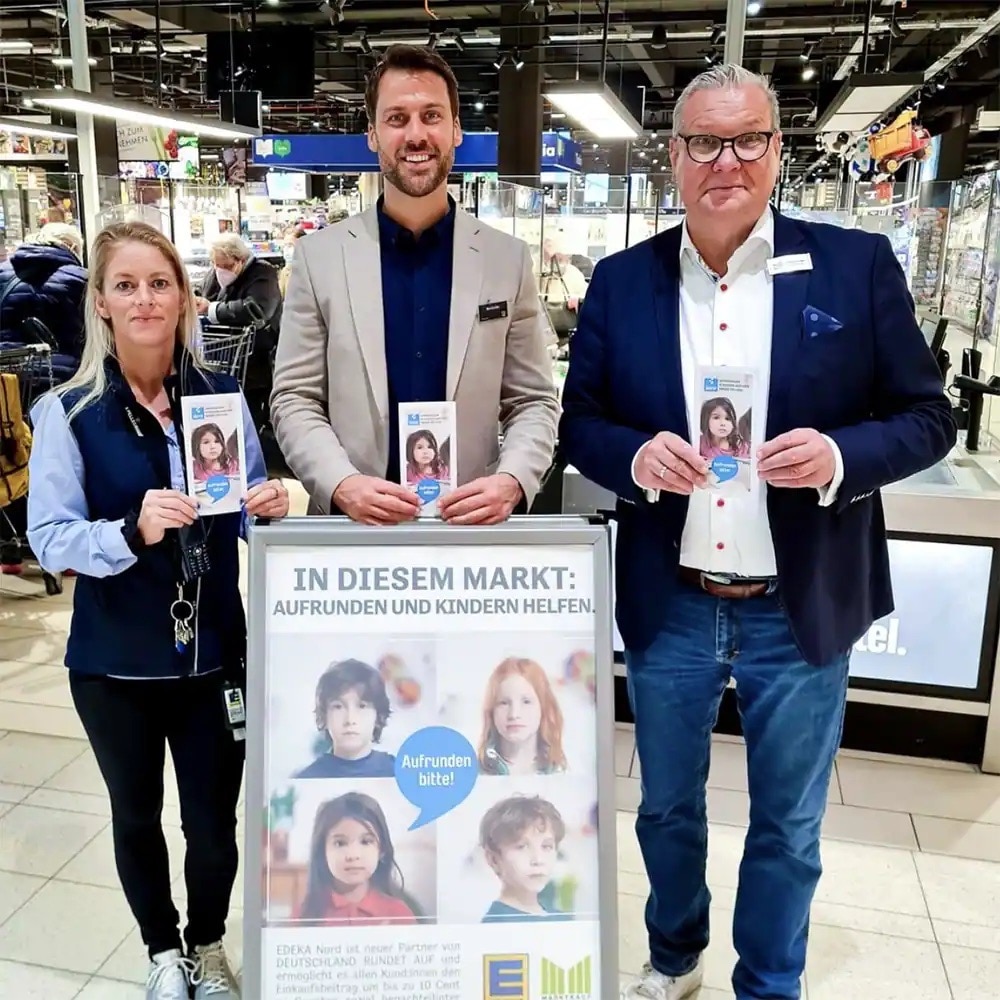 Drei Personen mit Flyern an einem Aufsteller mit Aufschrift: In diesem Markt aufrunden und Kindern helfen.