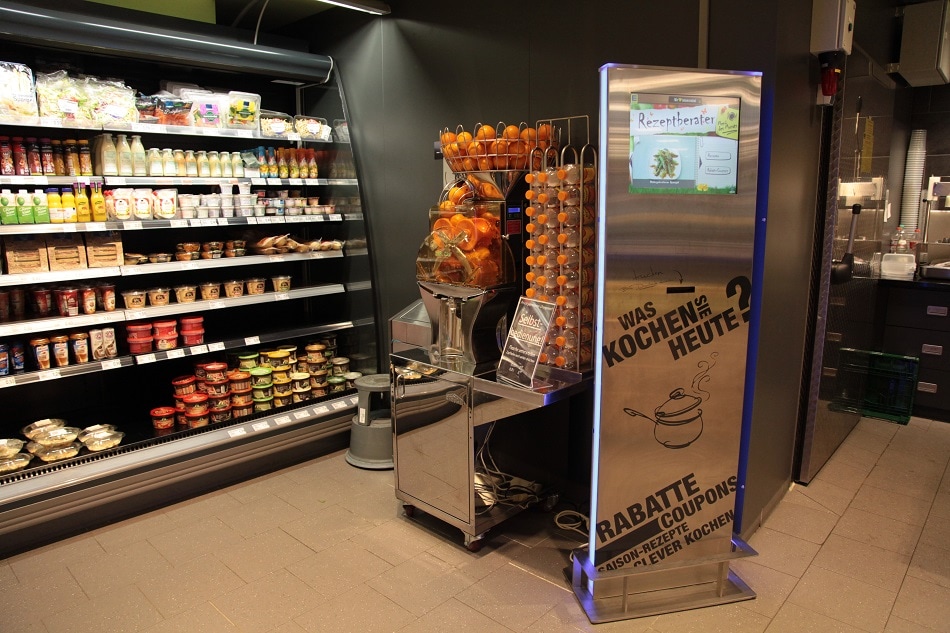 Neben einem Kühlregal steht eine Presse für Orangensaft sowie ein digitaler Rezeptberater.