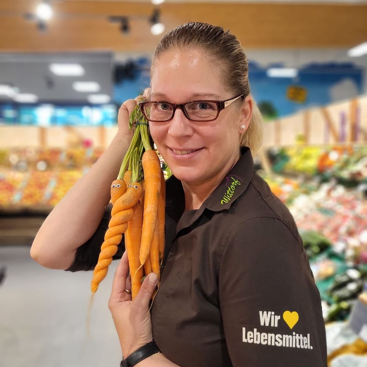 Eine Frau mit Brille und EDEKA-Hemd hält ein Bund Mohrrüben hoch. Auf zwei von ihnen sind Deko-Augen geklebt.