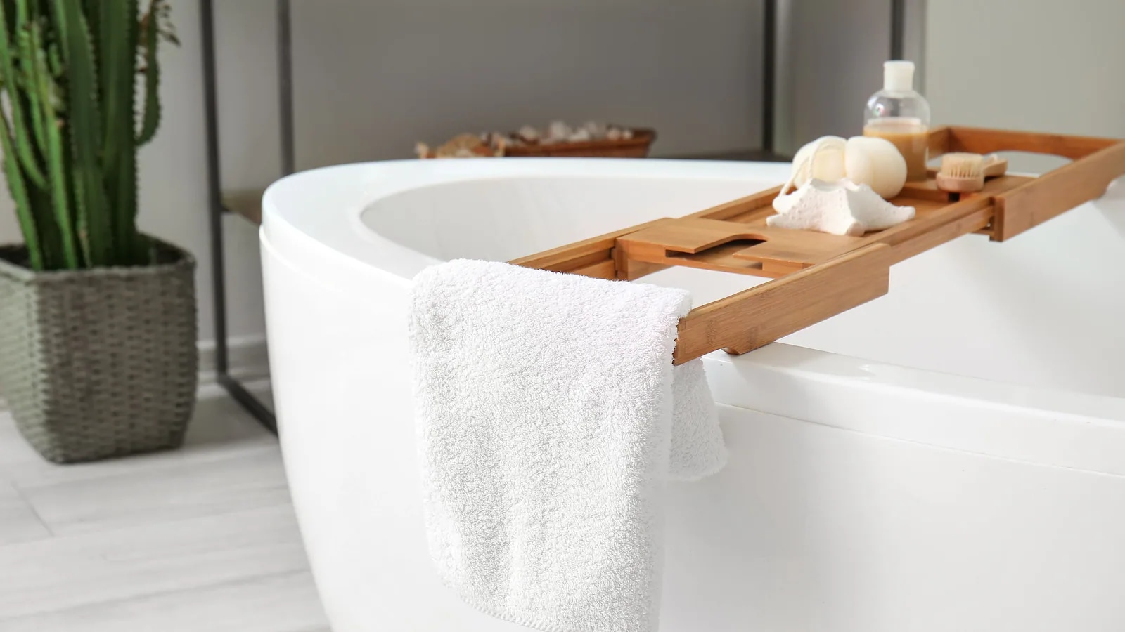 Freistehende weiße Badewanne mit einem Holz-Badewannenaufsatz, auf dem Pflegeprodukte liegen; ein weißes Handtuch hängt über dem Rand. Im Hintergrund steht ein Kaktus.