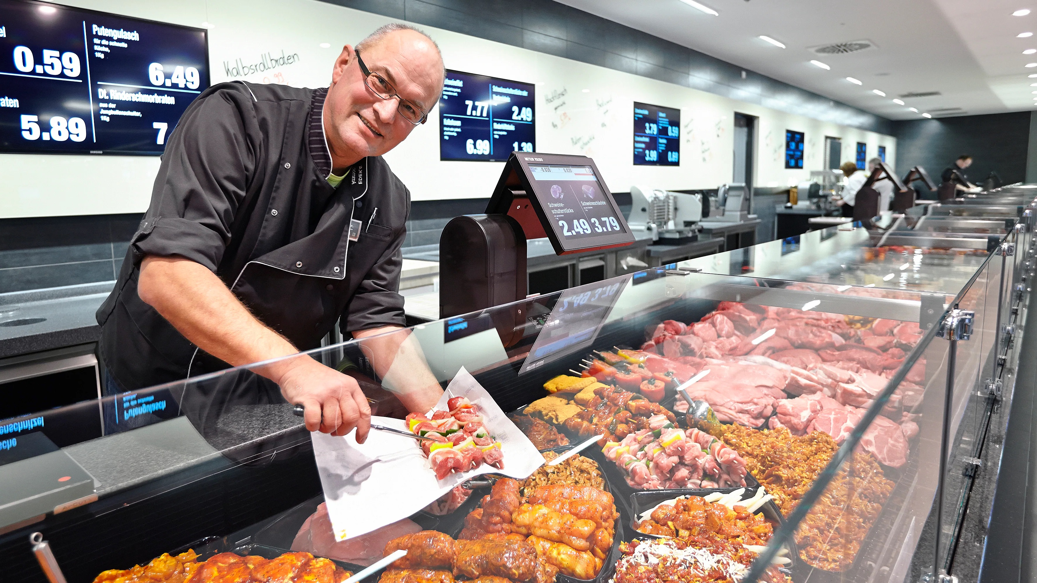 Blick entlang einer Bedientheke aus der ein lächelnder Mitarbeiter Fleischspieße entnimmt.