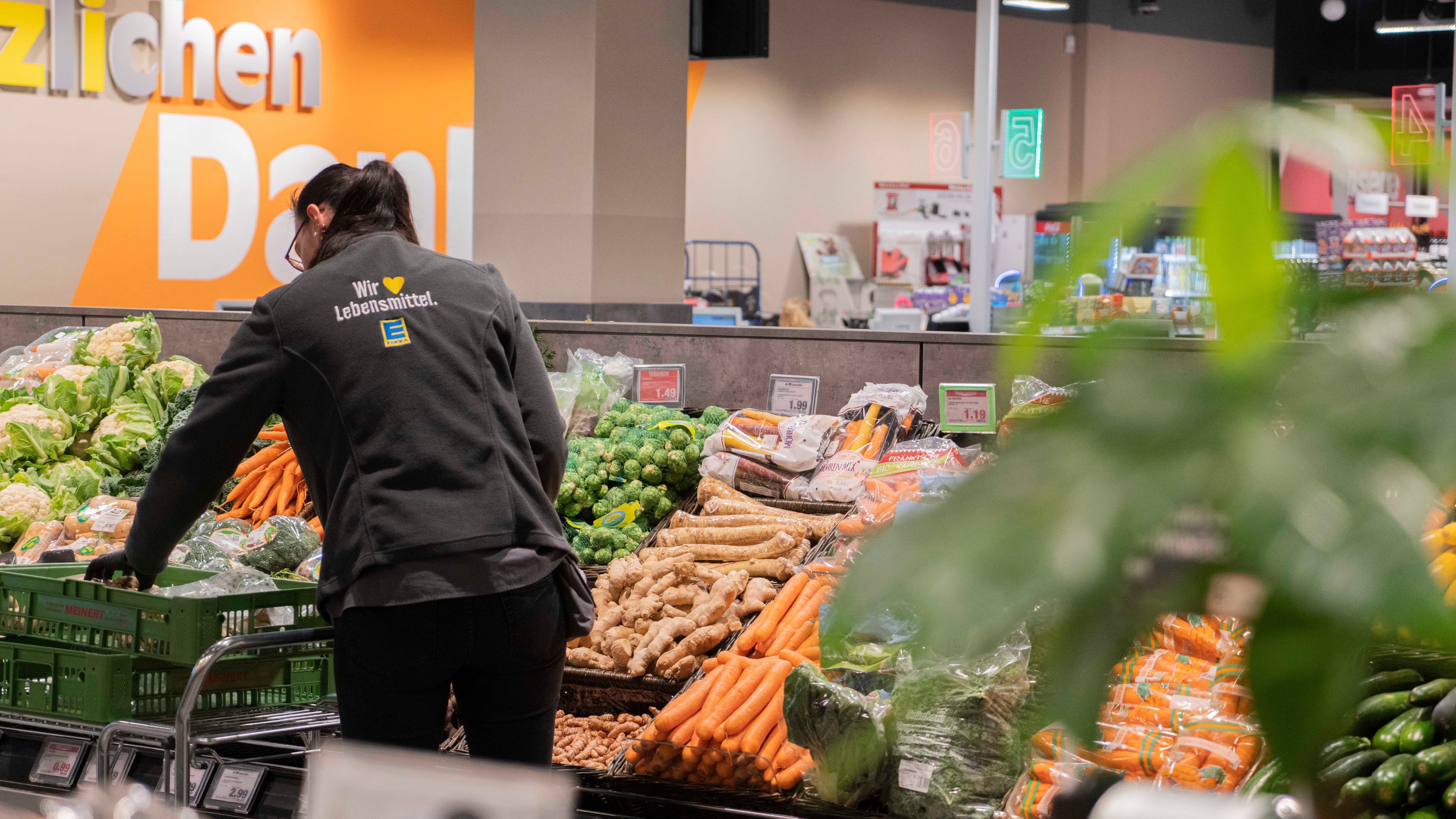 Eine Gemüsetheke mit verschiedenen Gemüsesorten zu dem eine Person die, die Theke mit neuem Gemüse bestückt.