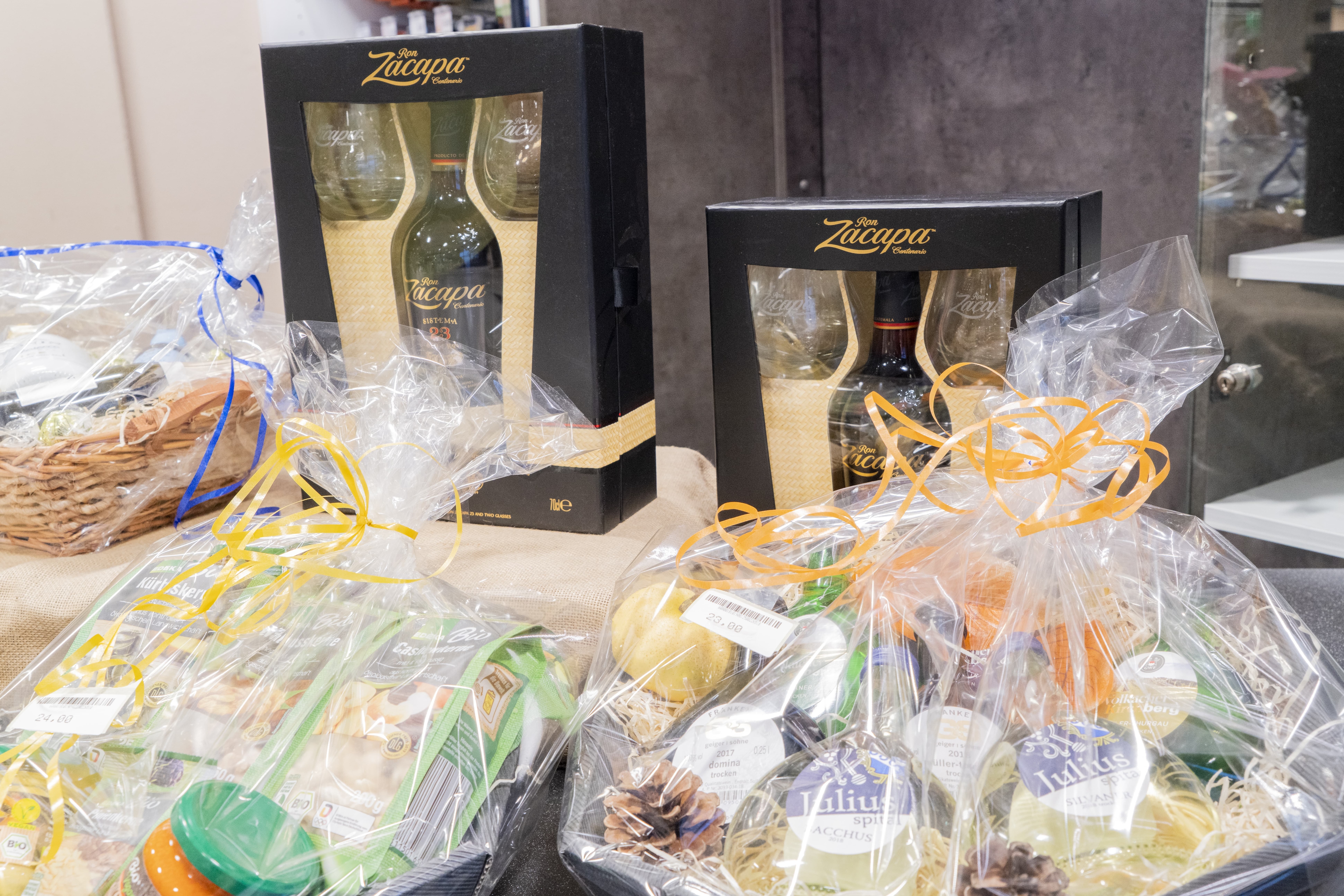 Drei Geschenkkörbe mit Delikatessen, verpackt in transparenter Folie und Geschenkband, im Hintergrund zwei Kartons mit Spirituosen und passenden Gläsern.