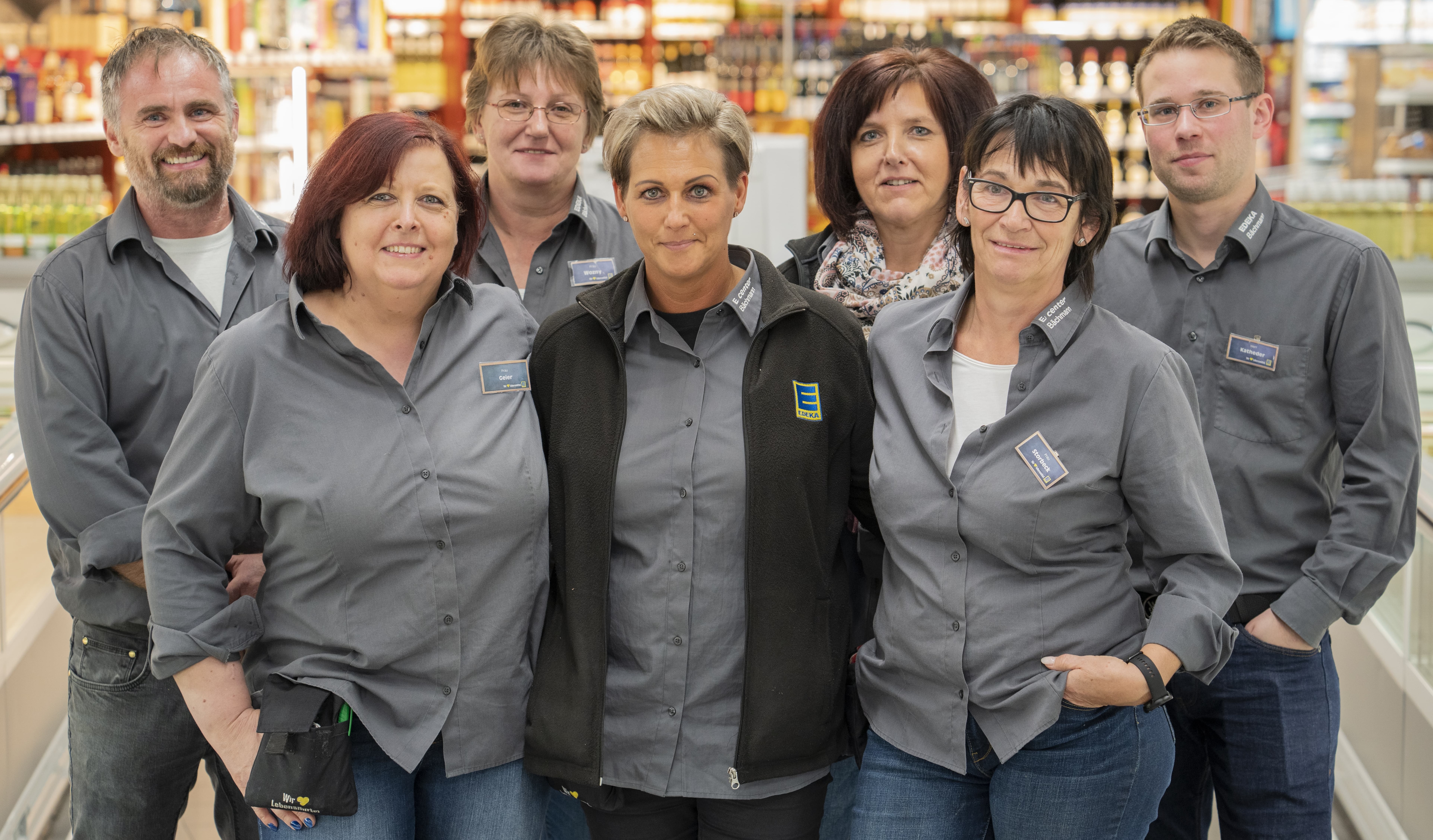 Mitarbeitende dieses EDEKA-Markts in grauer Arbeitskleidung, im Hintergrund verschwommen der Innenbereich des Markts.