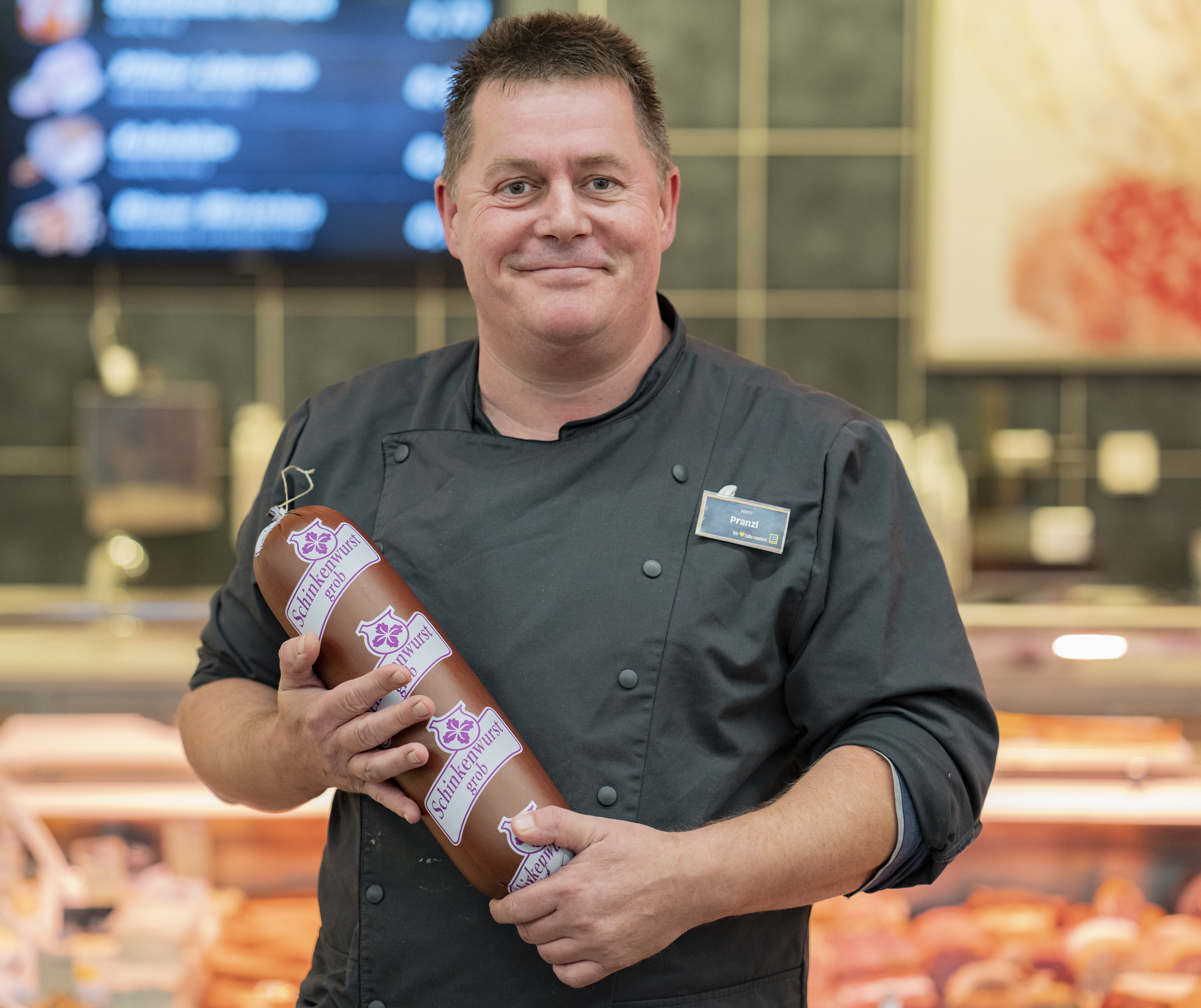 Ein Mitarbeiter lächelnd vor der Fleischtheke eines EDEKA-Markts. Er hält eine verpackte Wurstspezialität in den Händen.