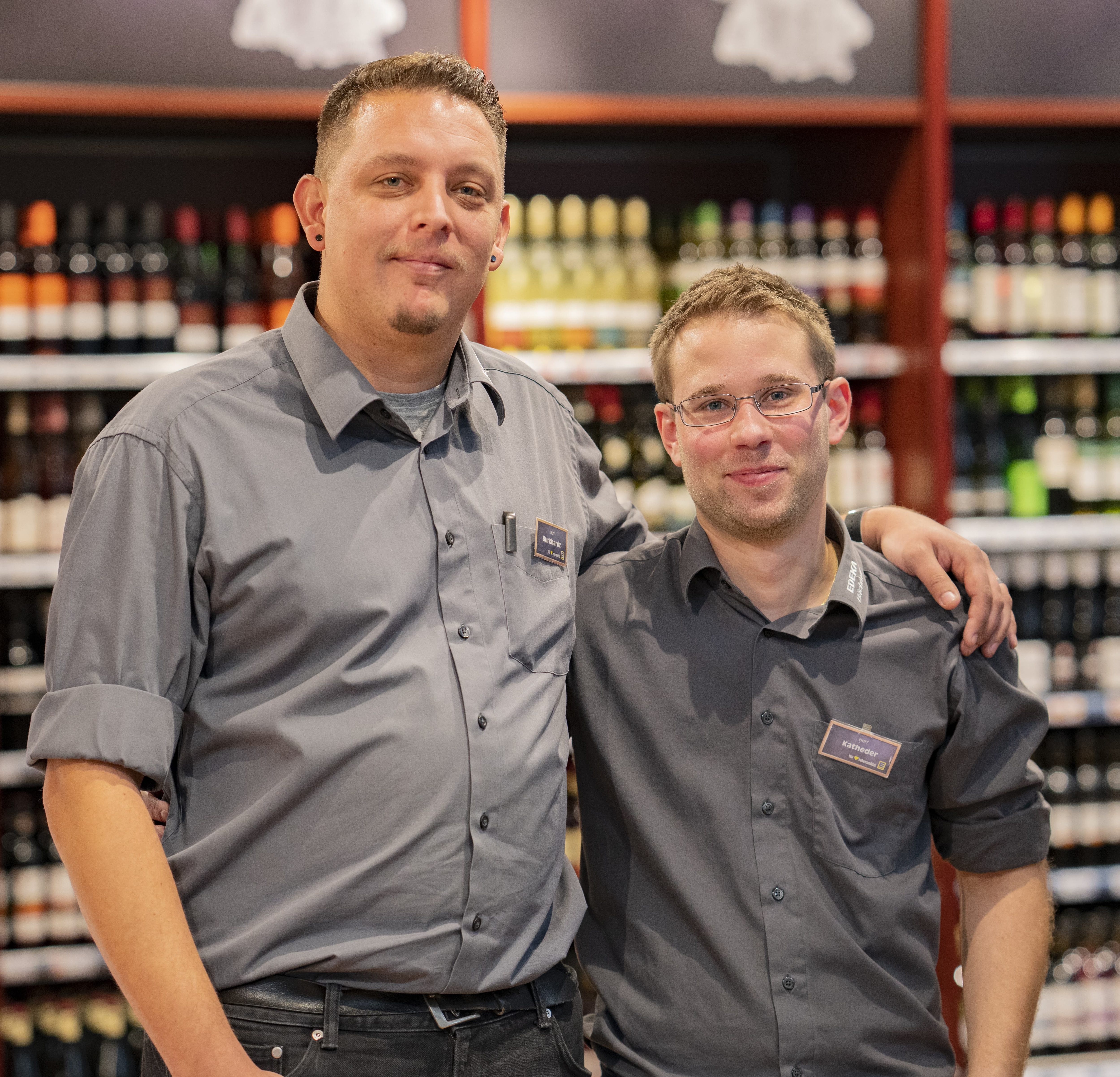 Zwei Mitarbeiter dieses EDEKA-Markts stehen Arm in Arm vor dem Alkoholsortiment des Markts und lächeln in die Kamera.