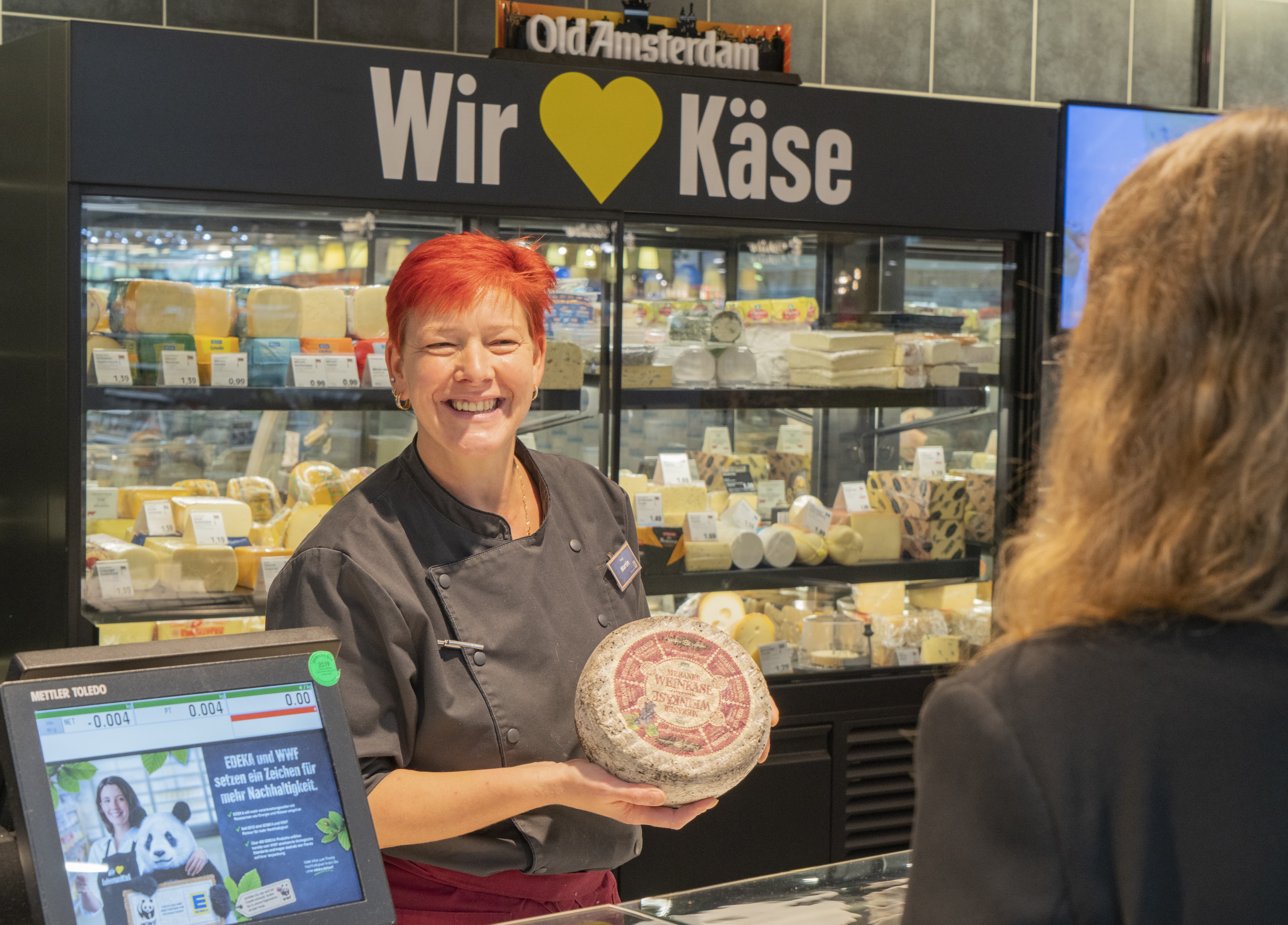 Eine Mitarbeiterin steht lächelnd hinter einer Käsetheke eines EDEKA-Markts. Sie hält ein Käserad in den Armen. Vor ihr steht eine Kundin.