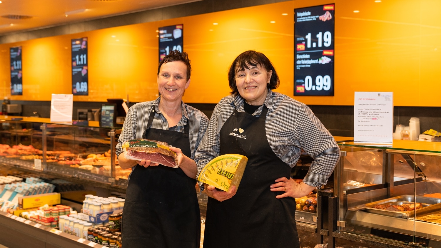 Zwei lächelnde Mitarbeiterinnen einer Bedientheke. Eine hält ein Stück Fleisch und die andere ein Viertel eines Käserads in den Händen.