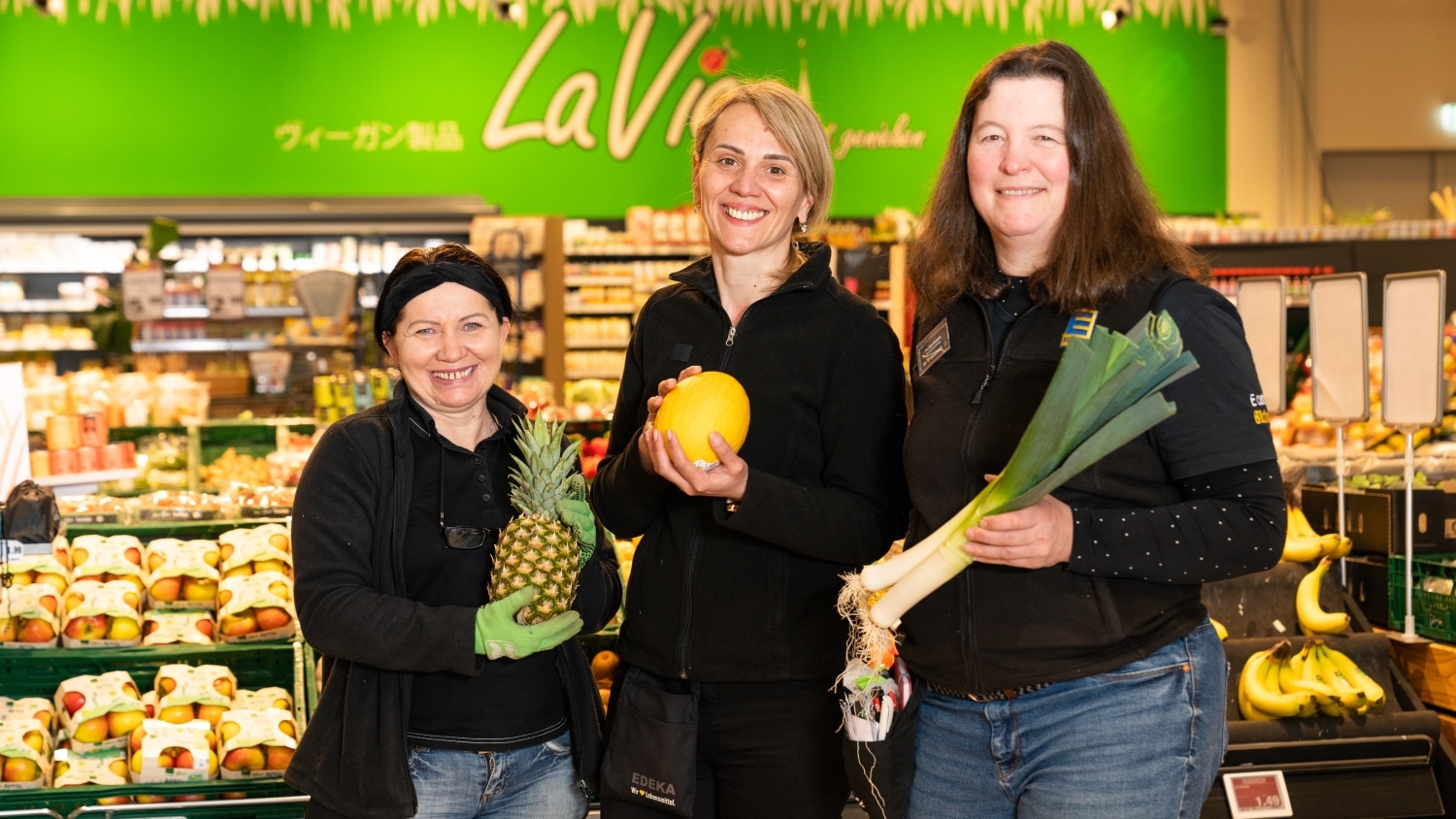 Drei lächelnde Mitarbeiterinnen halten verschiedene Obst- und Gemüsesorten in den Händen.
