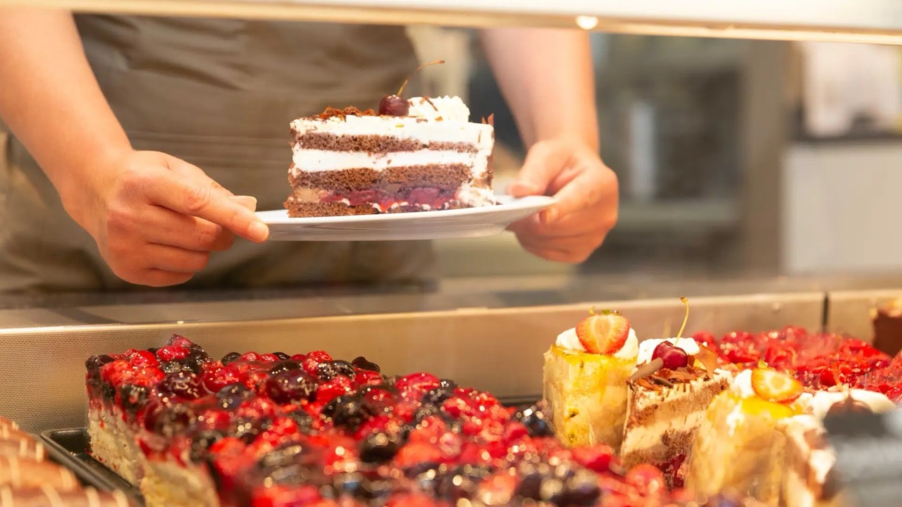 Zwei Hände nehmen einen Teller mit einem Stück Torte aus einer Theke.