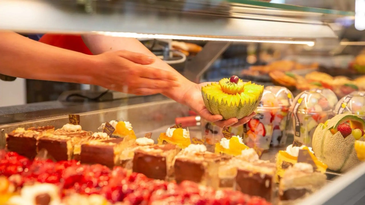 Eine Hand nimmt eine halbierte Melone aus einer Theke mit Kuchenstücken und abgepackten Obst.