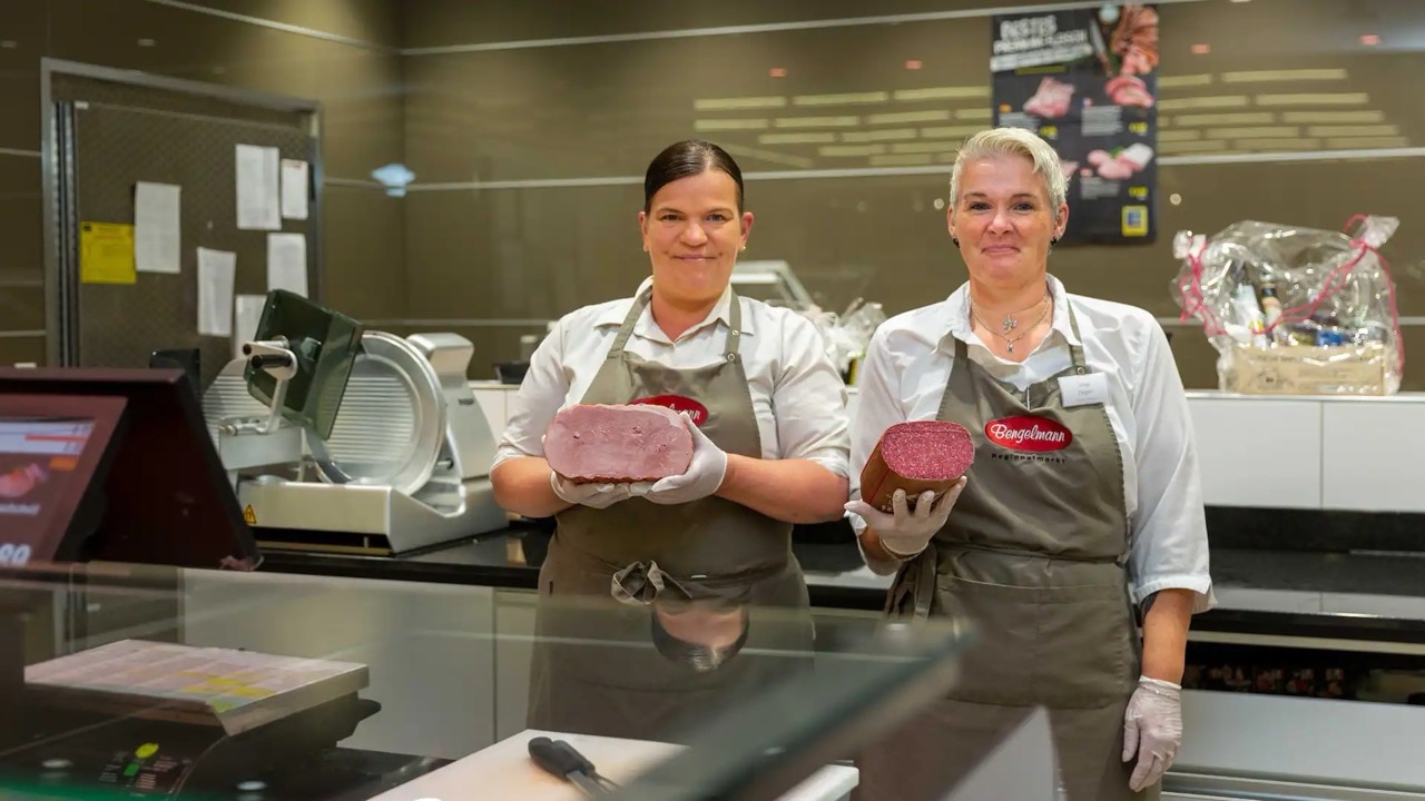 Zwei lächelnde Mitarbeiterinnen der Fleischtheke halten jeweils ein Stück Fleisch in den Händen.