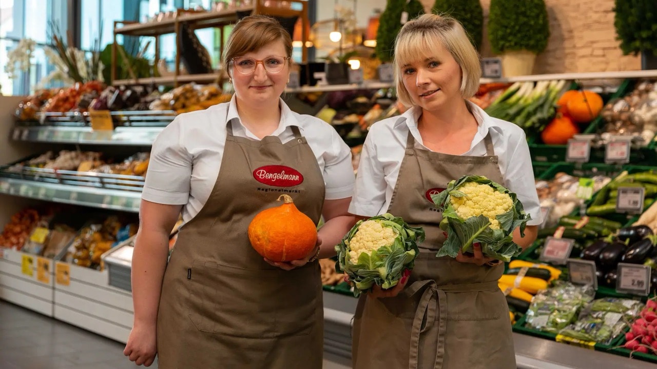 Zwei lächelnde Mitarbeiterinnen eines EDEKA-Marktes stehen in der Gemüseabteilung und halten verschiedene Gemüsesorten in den Händen.