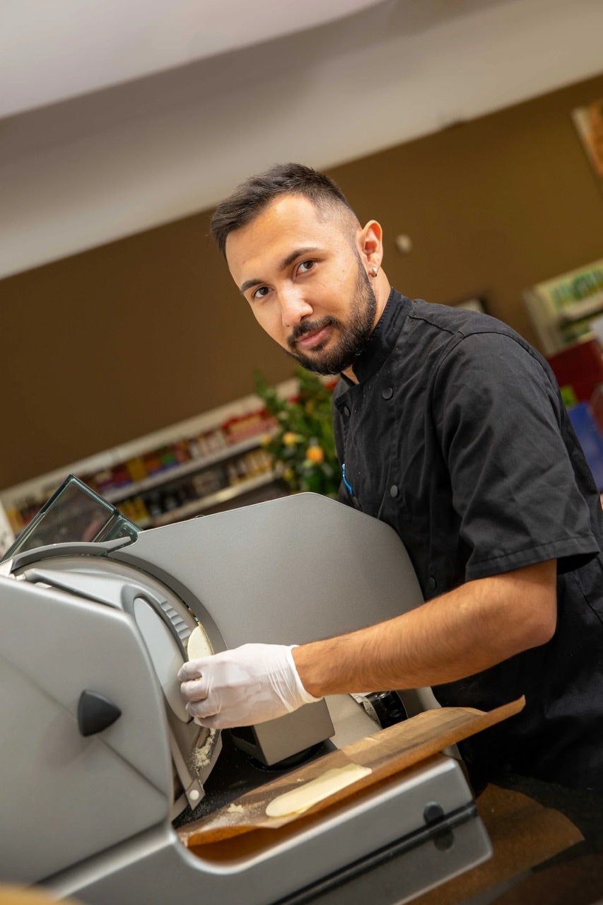 Ein lächelnder EDEKA-Mitarbeiter steht an einer Schneidemaschine und schneidet Käse.