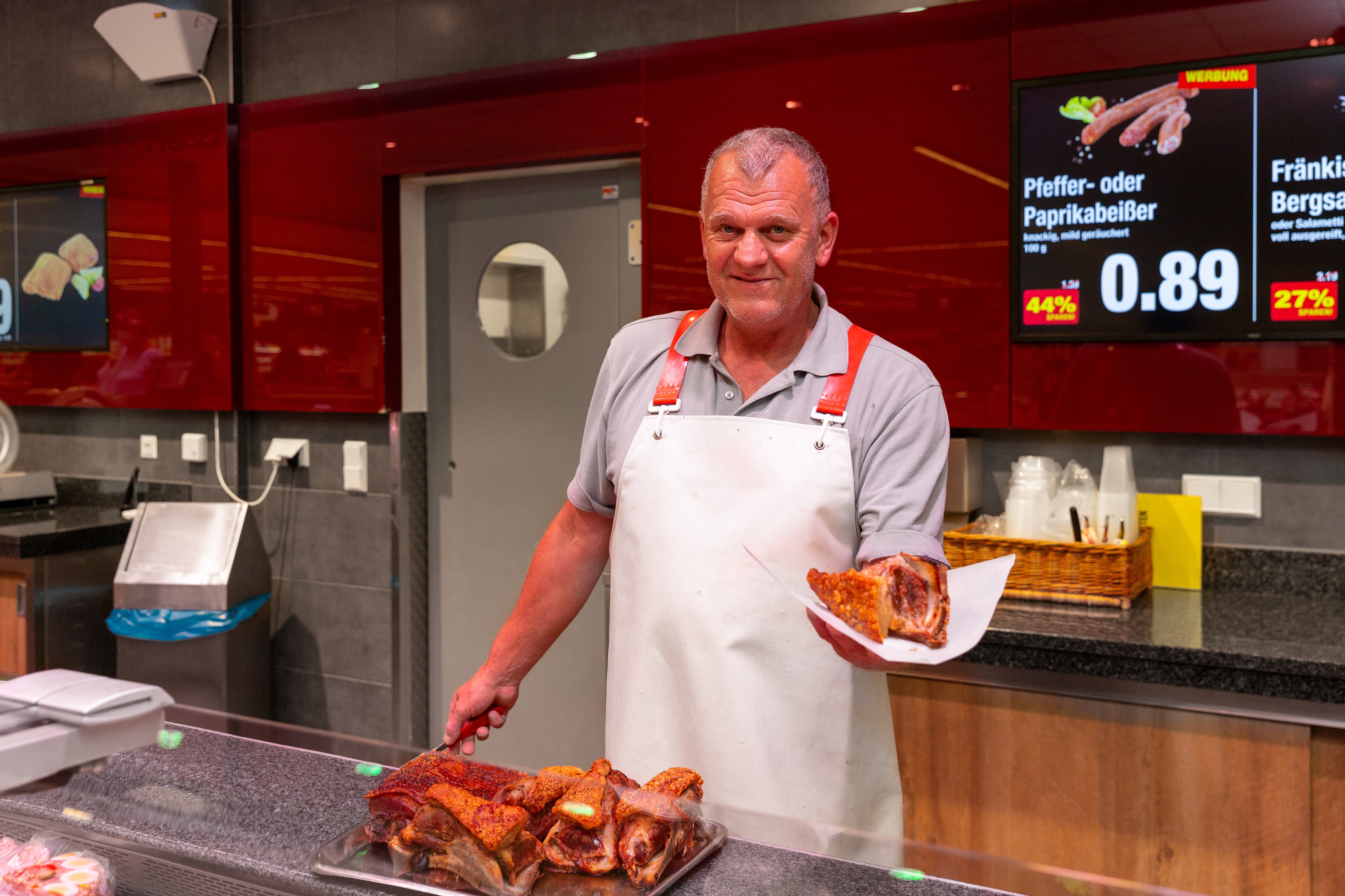 Ein Mitarbeiter der Fleischtheke präsentiert ein Stück Fleisch auf einem Stück Papier und lächelt in die Kamera.