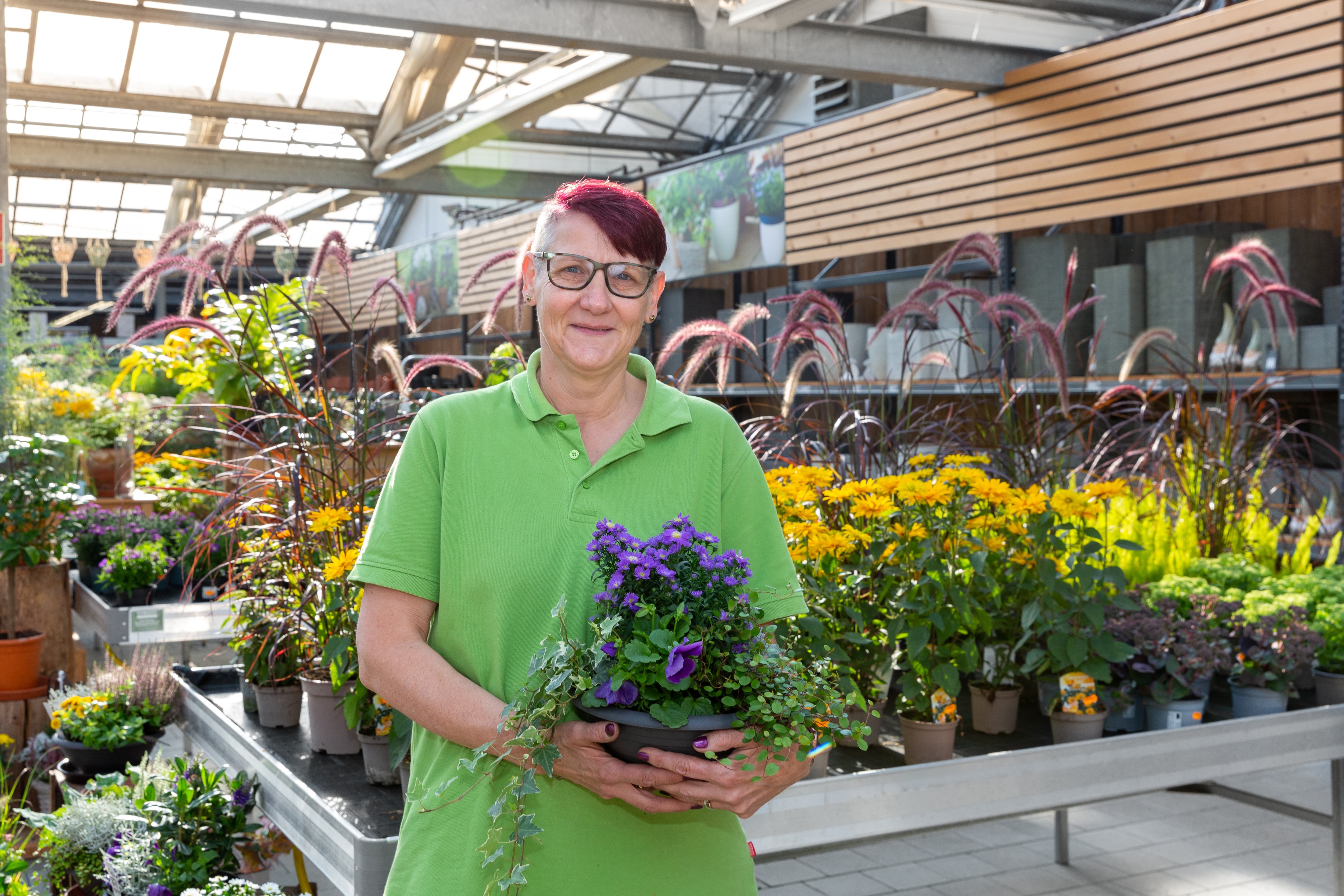 Eine lächelnde Mitarbeiterin steht im Gartencenter und hält eine Topfpflanze in den Händen.