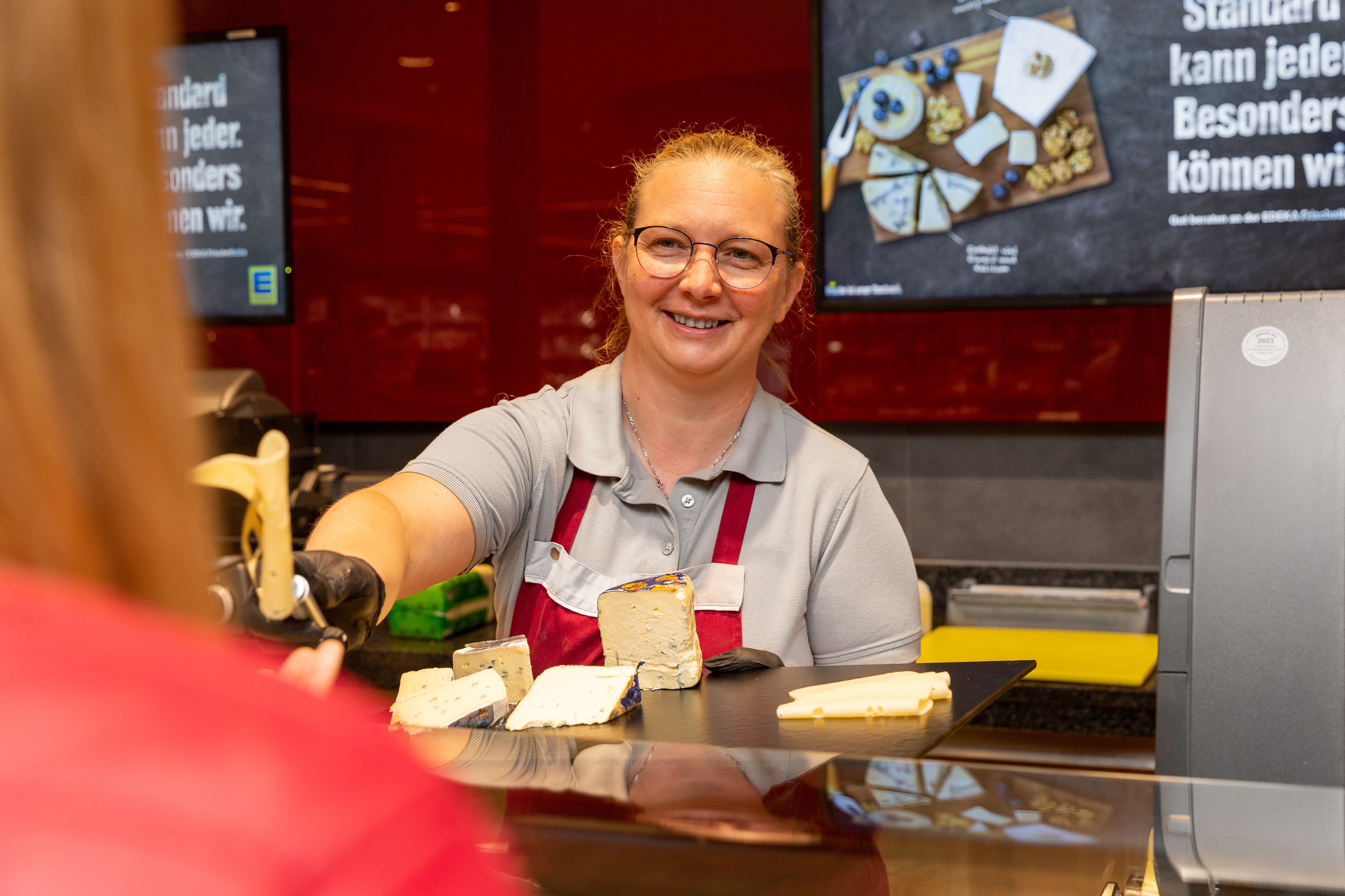 Eine Mitarbeiterin der Käsetheke reicht einer Kundin eine Scheibe Käse und lächelt dabei.